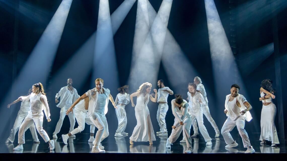 The greek gods dressed in white dancing under white beams of lights.