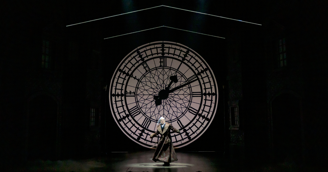 Scrooge in front of giant clock face.