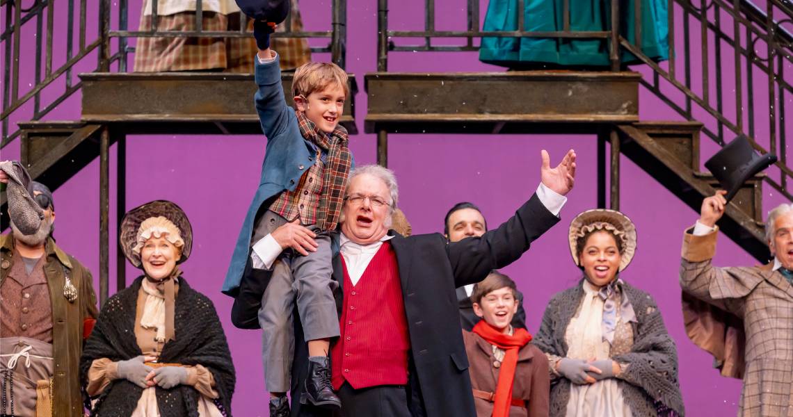 Hugo Verderame (Tiny Tim), Andrew May (Ebenezer Scrooge) and (background) the cast of A CHRISTMAS CAROL at Cincinnati Playhouse in the Park. Photo by Mikki Schaffner.